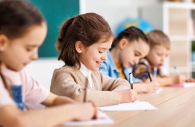Okulda sınav sırasında sınıf arkadaşlarının yanında otururken gülümseyen ve not defterine veri yazan pozitif küçük öğrenci.