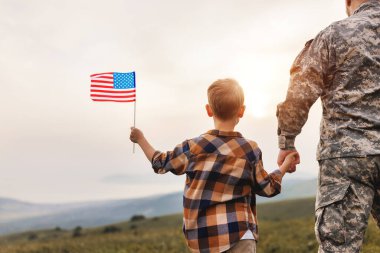 Amerikan bayrağıyla oğlunun elini tutan ve güneşli bir günde harika bir yaz manzarasının keyfini çıkaran asker babasının Amerikan ordusundan sonra oğluyla tekrar bir araya geldiği görüntüsü.