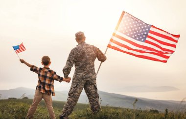 Amerikan bayrağıyla oğlunun elini tutan ve güneşli bir günde harika bir yaz manzarasının keyfini çıkaran asker babasının Amerikan ordusundan sonra oğluyla tekrar bir araya geldiği görüntüsü.