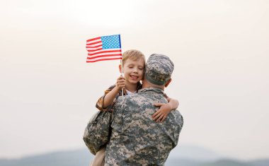 Amerikan bayrağını taşıyan mutlu küçük oğlan çocuğu askerî üniformalı babasına sarıldı. Amerikan ordusundan geri döndü. Yazın yeşil çayırda dururken erkek askerin ailesiyle tekrar bir araya geldiğini gördü.