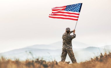 Gün batımında, bir dağın tepesinde Amerikan ordusu üniformalı bir erkek askerin sırtından ABD bayrağını sallarken.