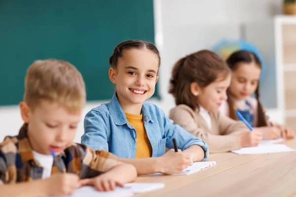 Positive little student smiling and writing down data into notebook while sitting at table near classmates during test at school