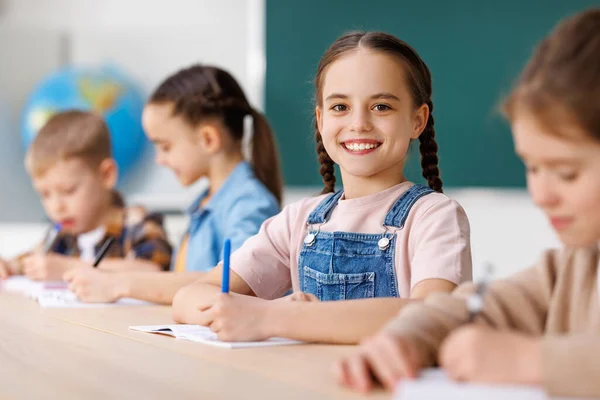Pozitív Kis Diák Mosolyog Írja Adatokat Noteszbe Miközben Asztalnál Közelében — Stock Fotó
