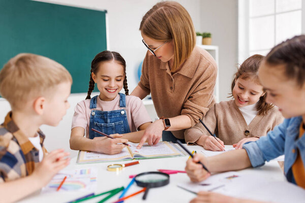 Smart woman teacher in  glasses helping students to to schoolwork during lesson in light classroom at school