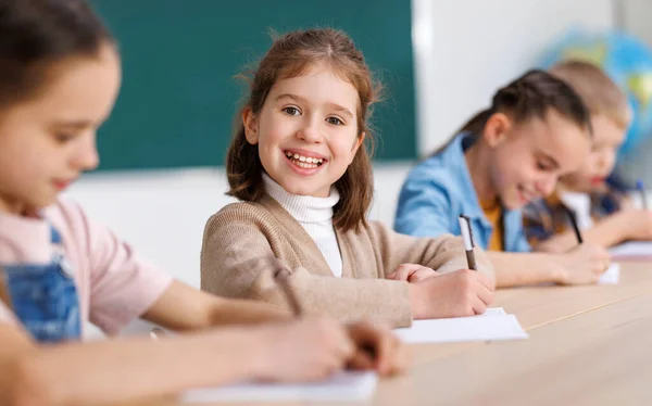 Positive little student smiling and writing down data into notebook while sitting at table near classmates during test at school