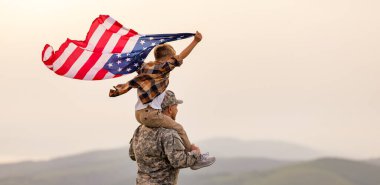 Amerikan bayrağını omuzlarında taşıyan mutlu bir çocuk taşıyan ve güneşli bir günde yaz manzarasının tadını çıkaran asker babası mutlu erkek asker baba, Amerikan ordusundan sonra oğluyla tekrar bir araya geldi.