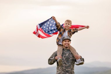 Amerikan bayrağını omuzlarında taşıyan mutlu bir çocuk taşıyan ve güneşli bir günde yaz manzarasının tadını çıkaran asker babası mutlu erkek asker baba, Amerikan ordusundan sonra oğluyla tekrar bir araya geldi.
