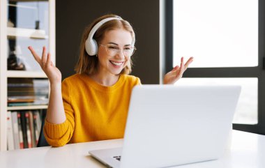 Gözlüklü neşeli genç bayan girişimci ofis masasında otururken gülümseyip el kol hareketi yapıyor ve online toplantı sırasında online iş arkadaşlarıyla konuşuyor.