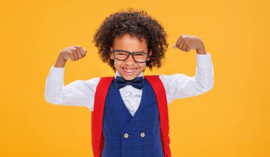 Afro-Amerikalı çocuk okul çocuğu güler ve güçlü kaslarını gösterir. Okul boyunca sarı arka planda eğlenirken.