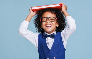 Gözlüklü, kameraya gülümseyen ve okul öğrencileri mavi arka planda eğlenirken kafasına kitap takan Afrikalı Amerikalı çocuk.