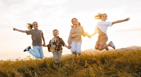Group of happy joyful school kids   jumping and holding hands in field on sunny spring day, excited children   boys and girls enjoying summer holidays in countryside