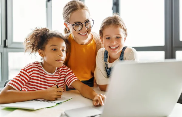 ハッピーな女性のチューターと2人の女子校生の子供たちが笑顔で 学校の日光浴教室でラップトップで教育ビデオを見ています — ストック写真