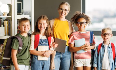Gündelik kıyafet ve gözlüklü mutlu kadın öğretmen ve güneşli derste kameraya bakan neşeli çocuklar.