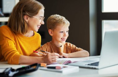 Gülümseyen öğretmen, anne ve okul çocuğu ödevlerini birlikte yaparken ve evde uzaktan çalışırken bilgisayarlarına göz atıyorlar.
