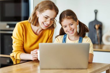 Annesiyle mutlu bir kız dizüstü bilgisayar kullanıyor ya da evdeki mutfakta online ders veriyor.
