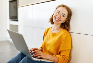 Neşeli genç bir kadın evinde dizüstü bilgisayar kullanıyor modern bir mutfakta yerde oturuyor.