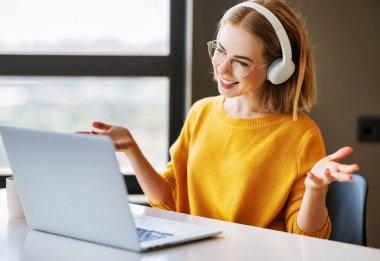 Gözlüklü neşeli genç bayan girişimci ofis masasında otururken gülümseyip el kol hareketi yapıyor ve online toplantı sırasında online iş arkadaşlarıyla konuşuyor.