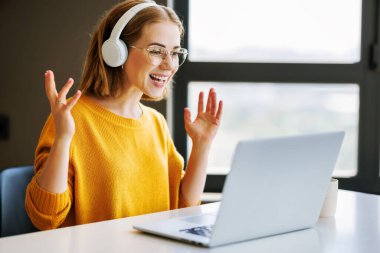 Gözlüklü neşeli genç bayan girişimci ofis masasında otururken gülümseyip el kol hareketi yapıyor ve online toplantı sırasında online iş arkadaşlarıyla konuşuyor.