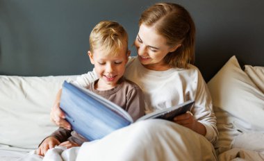 Neşeli aile: kadın ve erkek çocuk evde yatakta dinlenirken gülümseyip masal okuyorlar.