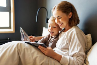 Neşeli aile: kadın ve erkek çocuk evde yatakta dinlenirken gülümseyip masal okuyorlar.