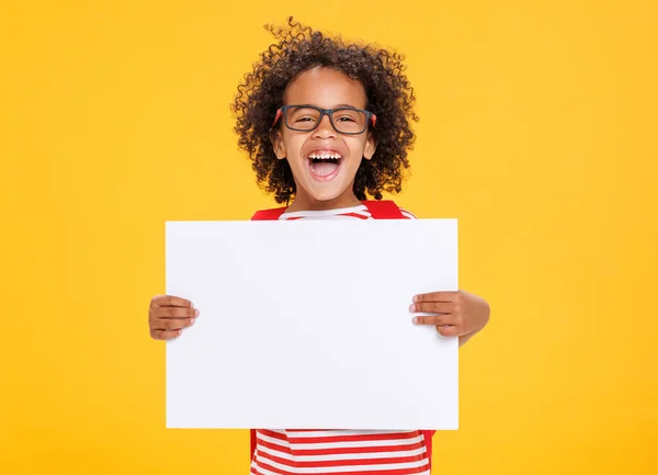 カメラのために笑い 黄色の背景に対して学校で楽しんでいる間に空のポスターを示すメガネの光栄なアフリカ系アメリカ人の少年 — ストック写真