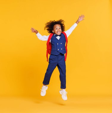 Uzun boylu, modaya uygun okul üniformalı Afro-Amerikan çocuk kollarını uzatıp sarı zemine atlıyor.