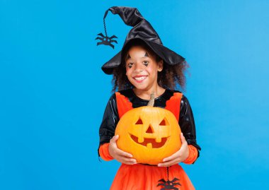 Balkabağı kostümü giymiş neşeli Afro-Amerikalı kız Cadılar Bayramı 'nı kutluyor ve parlak mavi arka planda gülüyor.