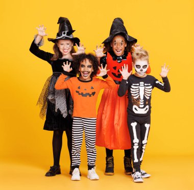 Happy Halloween! Cheerful kids in carnival costumes and makeup make a terrible gesture on bright colored yellow background clipart