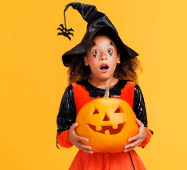 Sarı arka planda Cadılar Bayramı 'nı kutlarken elinde balkabağı ve çığlık atan kıvırcık saçlı mutlu Afrikalı Amerikalı kız.