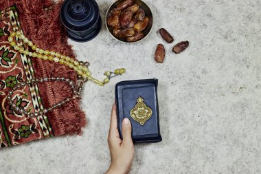 lantern and Holy Quran with Arabic calligraphy meaning of Al Quran and prayer mat , rosary muslim and bowl dates fruits and woman hands holding a holy book