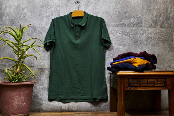 Front side of male green cotton t-shirt hanging on gray grunge marble wall with cactus pot and stacked shirts. T-shirt without print and copy space for your text on right side