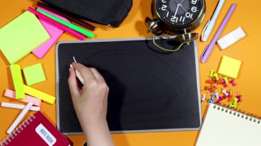 Turuncu arka planda okul malzemeleri ve okula dönen yazı tahtasında yazan bir kadın. 