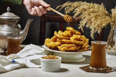 Griwech crunchy pastries , an Algerian dessert made with dough, fried and then coated with syrup and sesame seeds and woman holding  honey spoon  clipart