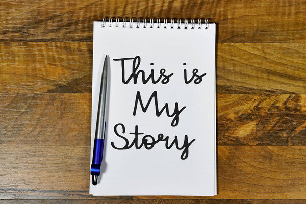 spiral notebook with the phrase "This is My Story" written in bold, cursive lettering, accompanied by a silver and blue pen, placed on a textured wooden surface. A personal and reflective composition