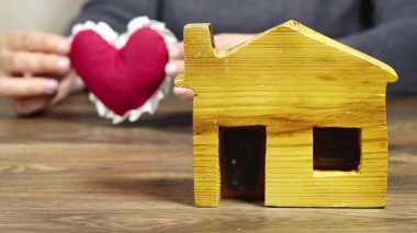 Ahşap bir ev figürü ve peluş kırmızı bir kalbi tutan bir el sevgiyi, sıcaklığı ve ev ve aile kavramının bağlantısını taşır.