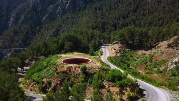 緑の斜面の端に位置する丸い形をした赤い湖の航空眺め クリップ 森林化された山々と珍しい自然現象 — ストック動画