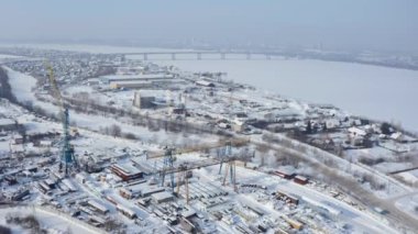 Şehrin karla kaplı sanayi bölgesinin havadan görünüşü. Şarjör. Beyaz kış şehrinin üzerinde nehir ve köprüyle uçuyor.