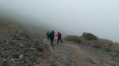 Bir grup aktif turist sis içinde dağ yolunda yürüyor. Şarjör. Çubuklu turistler bulutlu bir günde yokuş yukarı giderler. Dağda yoğun sis bulutlu bir günde turistlerle. 