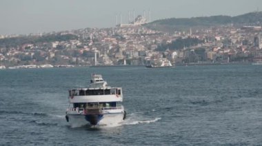 Limandaki mavi sularda beyaz yat kıyıdan yavaşça hareket ediyor. Başla. Arka planda dağlık bölgede yer alan şehir