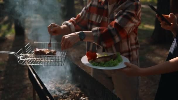森の中の自然のピクニック 煙でキャンプファイヤーの上のBbq ストック映像 おいしい鶏を野菜で女性の皿に入れる男 — ストック動画
