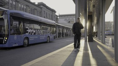 Sırbistan, Belgrad - 12 Nisan 2023: Genç adam otobüs durağında telefonla konuşuyor. Başla. Genç adam güneşli bir günde otobüs durağında otobüs bekliyor. Şehir yollarında modern elektrik otobüsleri.