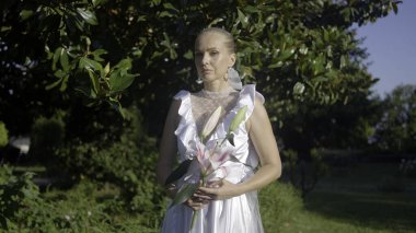 Beyaz elbiseli genç bir gelinin portresi. Elinde zambak çiçeği var. Başla. Arkasında yeşil ağaçlar olan güzel bir gelinin video çekimi.