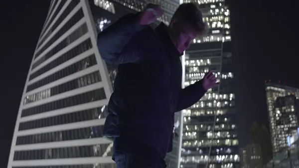 Man dances on background of skyscrapers. Action. Young man is dancing coolly on background of night city. Guy dances in modern style at skyscrapers at night.