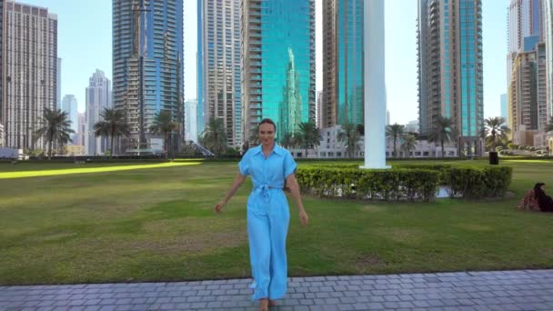 穿着蓝色夏装的快乐女人在城市公园里散步 背景上的绿色草地和摩天大楼 — 图库视频影像