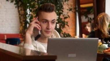 Genç adam kafede dizüstü bilgisayarla konuşuyor. Stok görüntüleri. Genç adam dizüstü bilgisayarla çalışırken telefonla konuşuyor. Adam kafede dizüstü bilgisayarla çalışırken iş çağrılarına cevap veriyor..