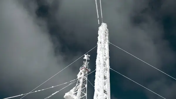 Snow-covered radio tower on background sky. Clip. Frozen structure of electric tower in winter. Radio towers covered with snow on sunny winter day.