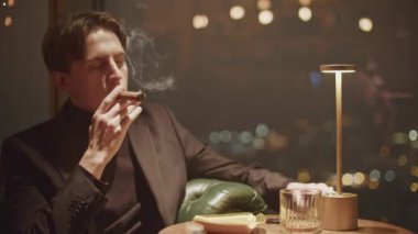 Siyah elbiseli genç adam karanlık bir restoranda puro içiyor. Medya. Düşünceli adam duman üflüyor. 