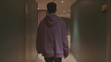 Oteldeki büyük boy pembe kapüşonlu genç adam. Borsa klipsi. Adam uzun koridordaki oda kapılarında yürüyor.