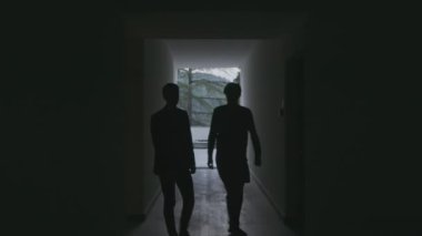 İki erkek siluet karanlık, uzun otel koridorunda yürüyor. Borsa klipsi. Siyah salonda erkek figürleri
