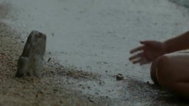 Kıyıda suyla oynayan çocuğun yakın çekimi. Yaratıcı. Kıyıda oturan çocuk su sıçratıyor. Çocuk dalgalarla su sıçratıyor..